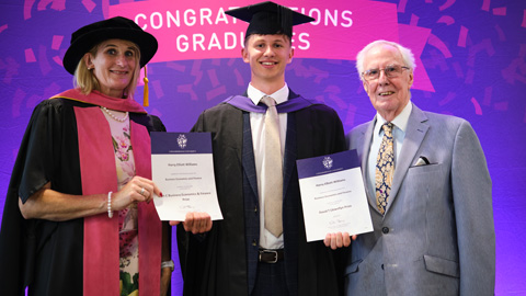 Three people at a graduation ceremony; two in academic regalia holding certificates, one in formal attire.